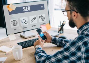 handsome young seo manager using smartphone and computer at workplace